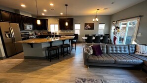A modern kitchen with dark cabinets, a granite countertop, and stainless steel appliances. There is a large island with stools, and a leather sofa in the living area. - Contemporary Family & Business Friendly House with Pool (Las Vegas)