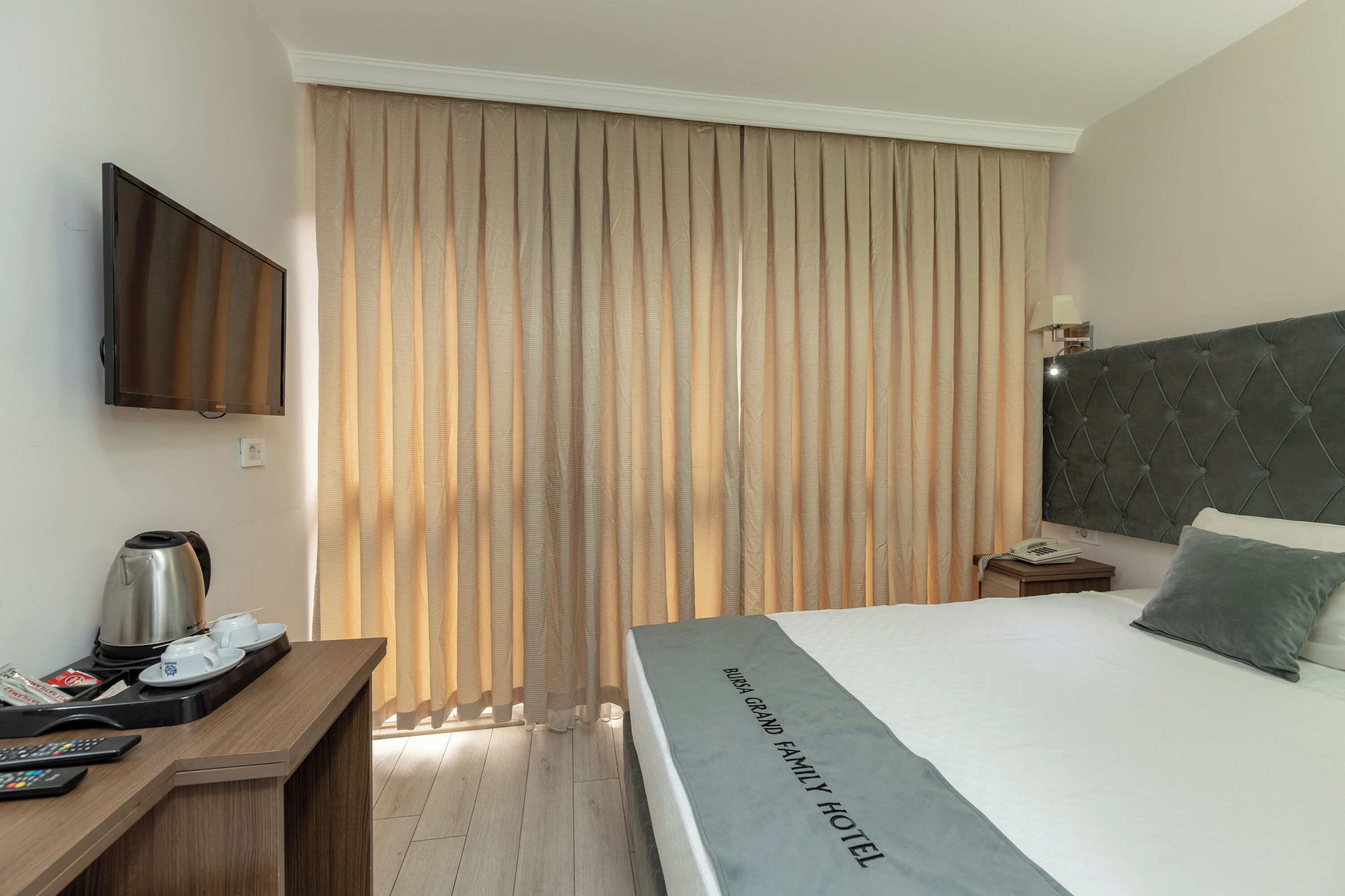 Quarto de hotel com cama, televisão na parede, mesa de cabeceira com abajur e janela com persianas verticais.