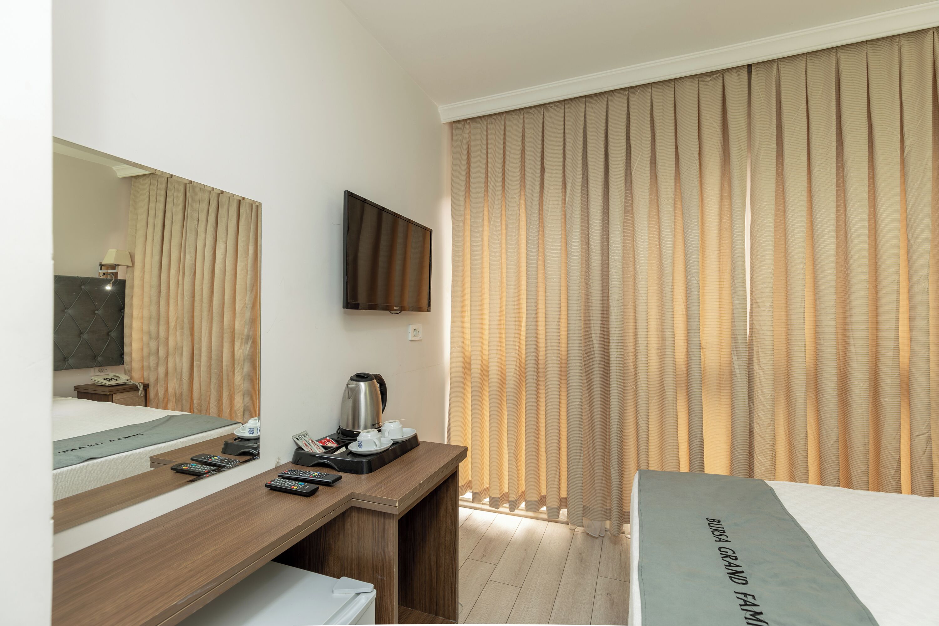 Een hotelkamer met een bed, een bureau met een flatscreen televisie, een koffiezetapparaat en een groot raam met verticale jaloezieën.