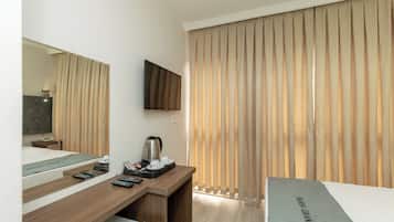 Een hotelkamer met een bed, een bureau met een flatscreen televisie, een koffiezetapparaat en een groot raam met verticale jaloezieën.
