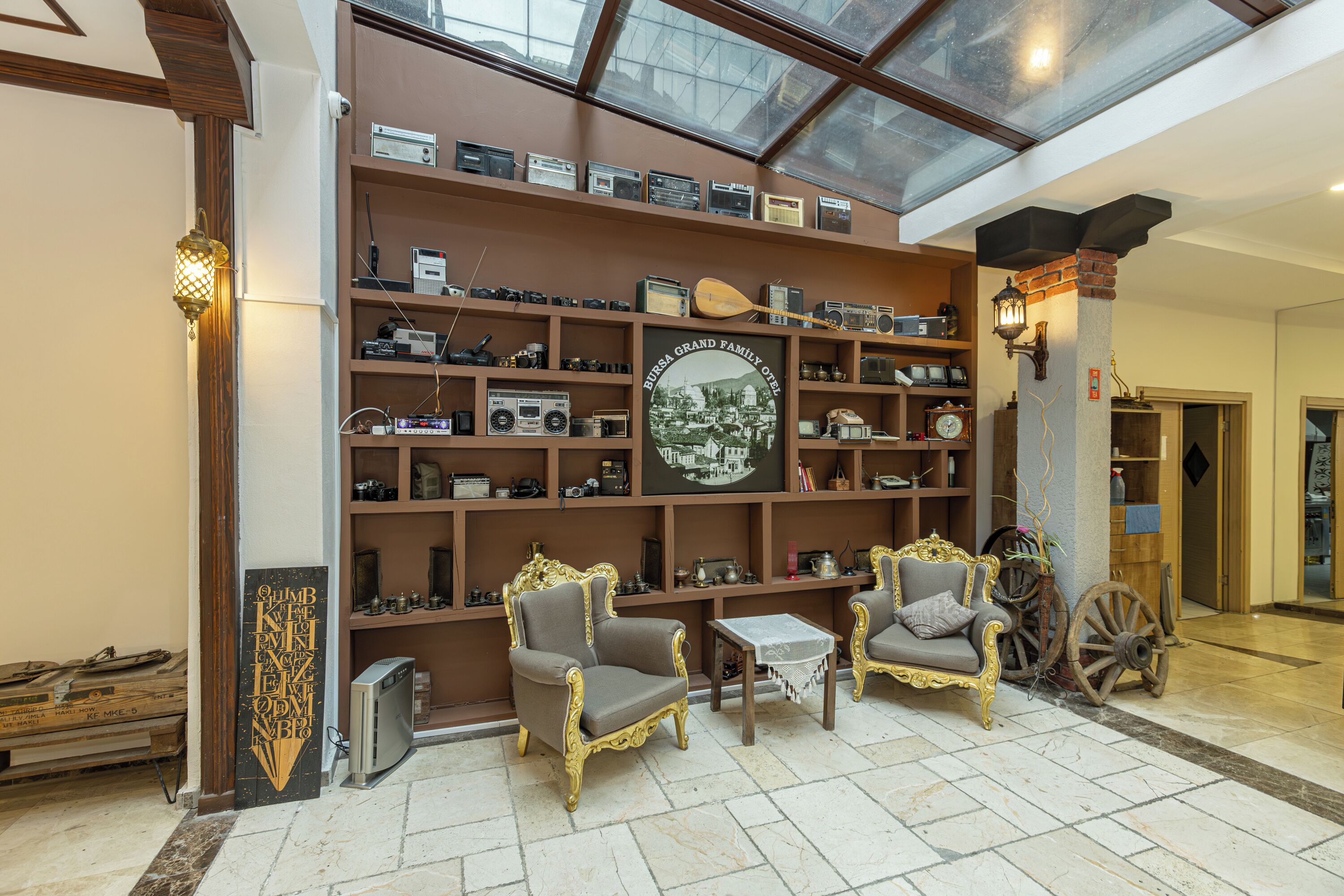 A lobby with a high ceiling, a glass roof, and a wall adorned with vintage radios and a large family photo. There are two ornate armchairs and a small table.