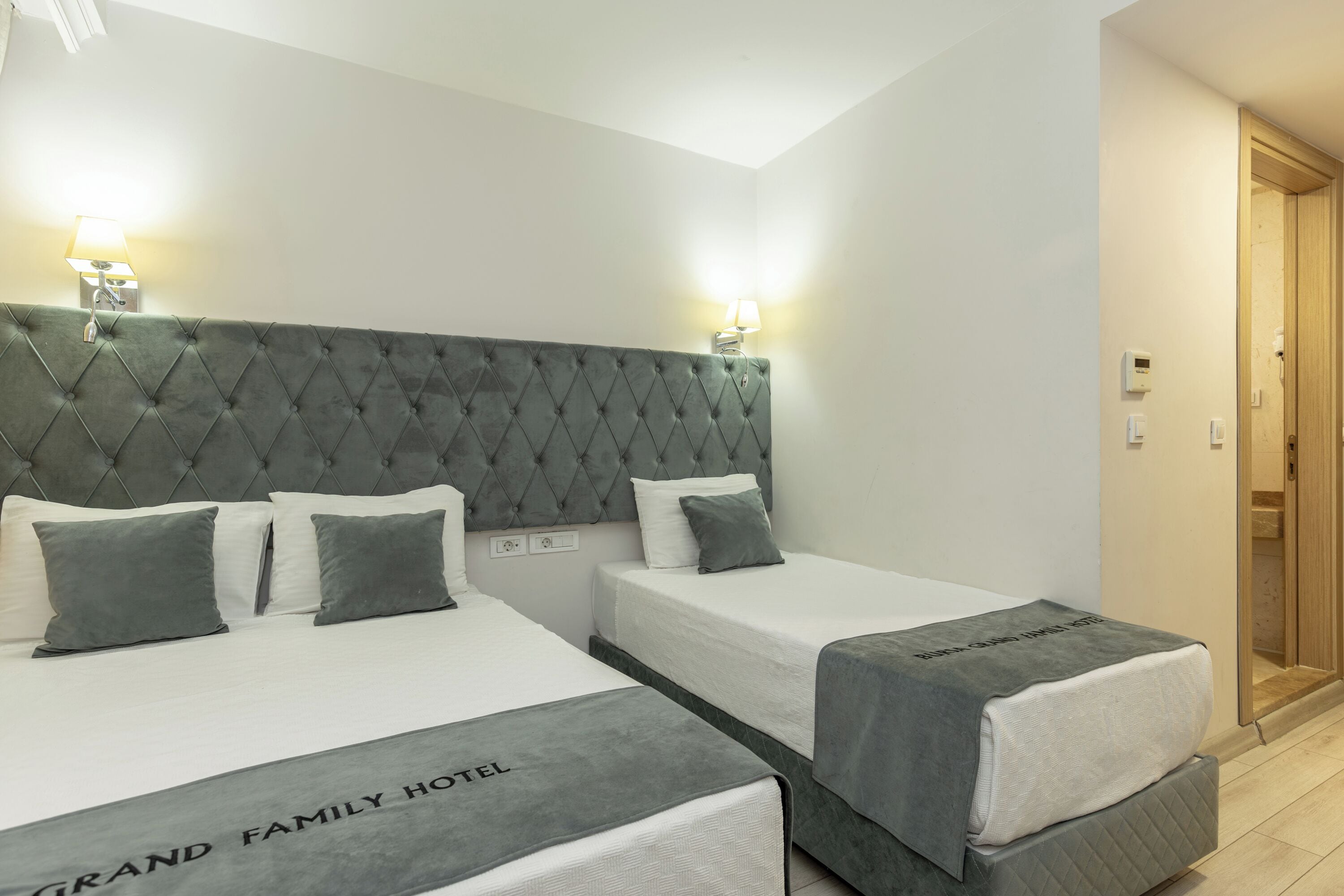 Een hotelkamer met twee bedden, een gestuwd hoofdeinde, nachtlampjes en een bordje met de tekst 'GRAND FAMILY HOTEL'.