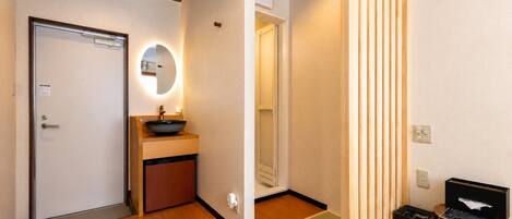 A hotel room with a wooden floor, a sink with a round mirror, a door, and a small trash bin.