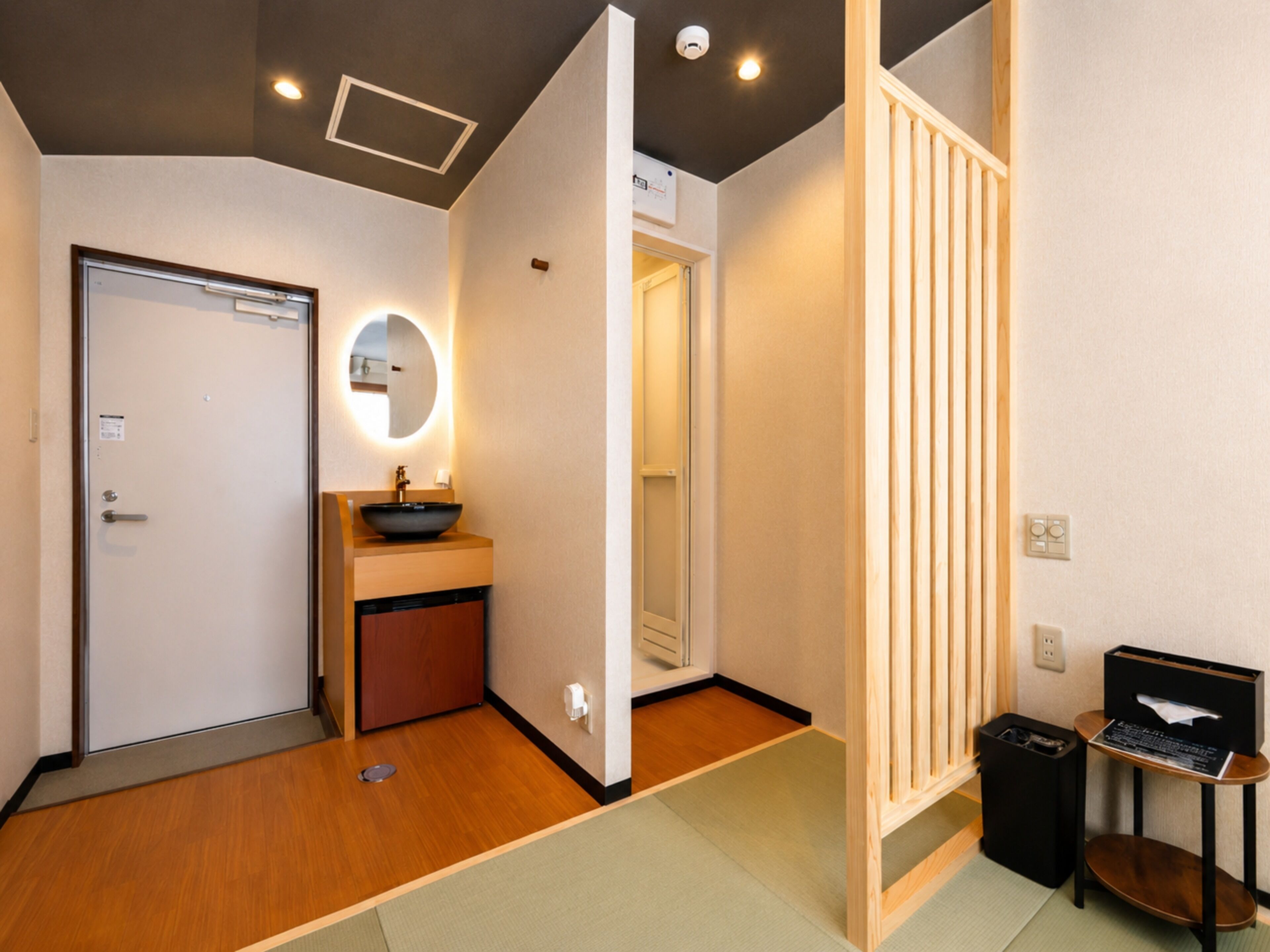 Hotelkamer met houten vloer, ronde spiegel boven een wastafel, een deur met een handgreep en een kleine prullenbak.