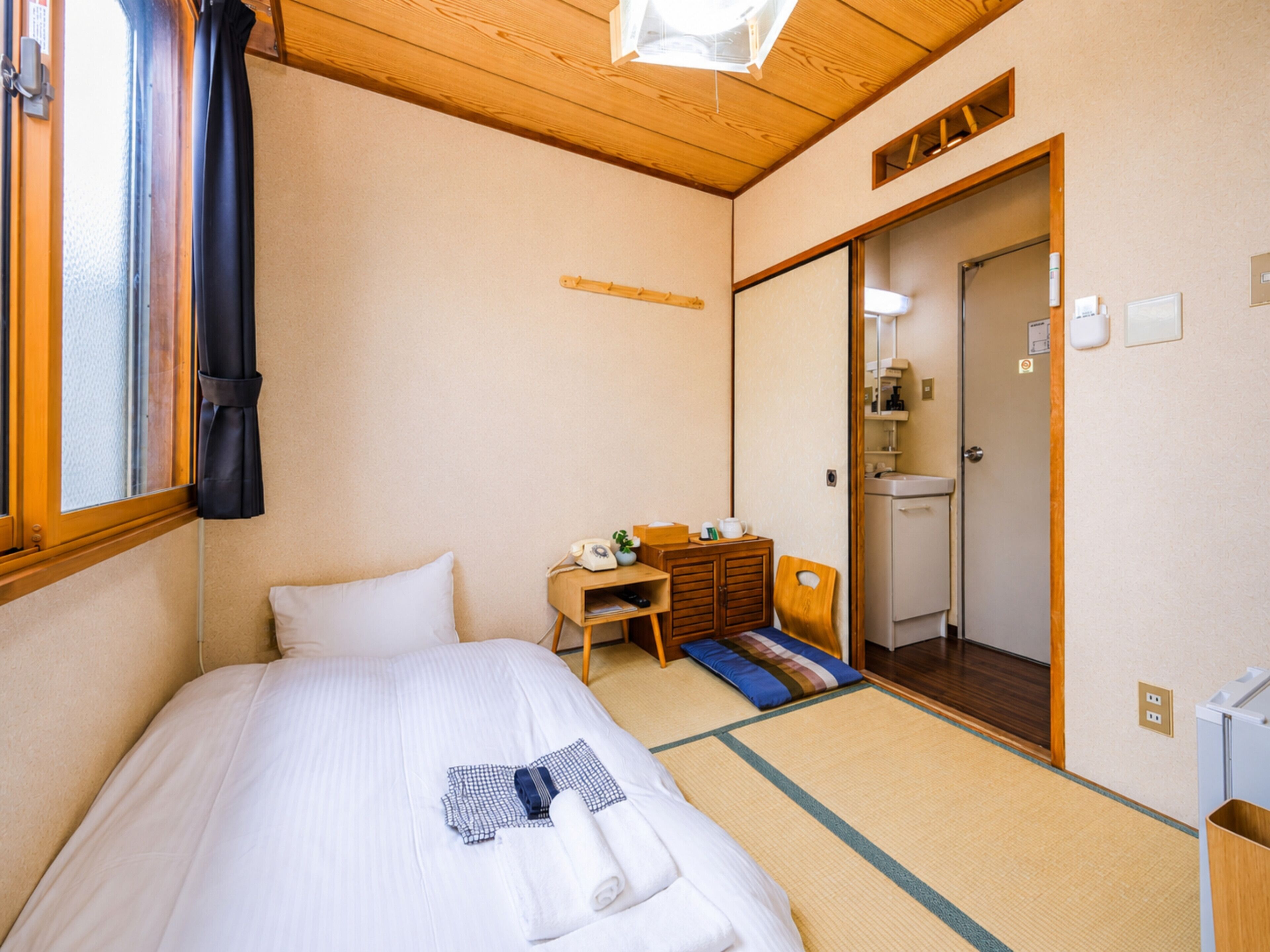 A hotel room with a bed, a desk, a chair, a small cabinet, a mirror, and a door leading to a bathroom.