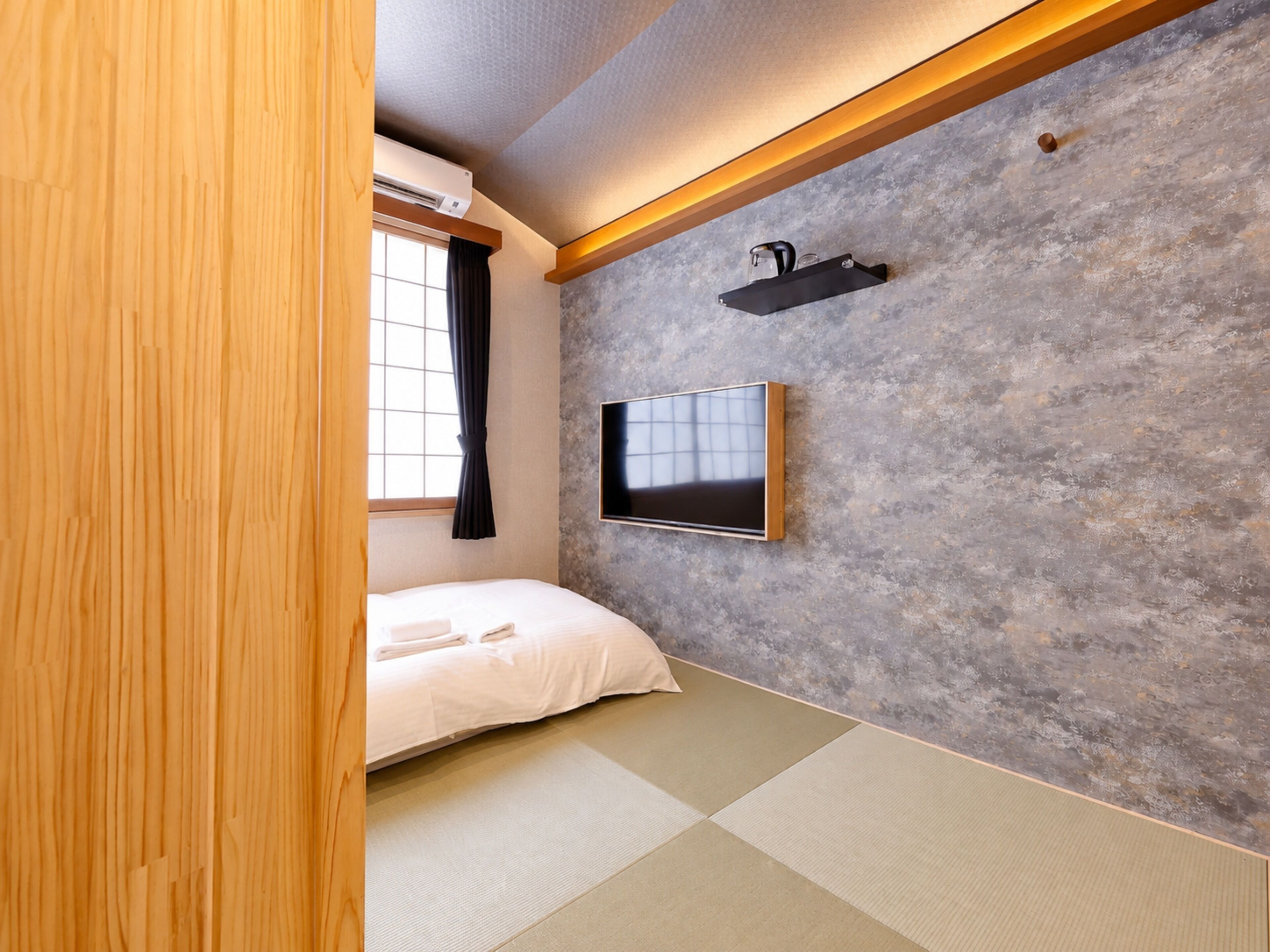 Een kamer met tatami vloerbedekking, een bed, een bevestigde televisie en een raam met gordijnen.