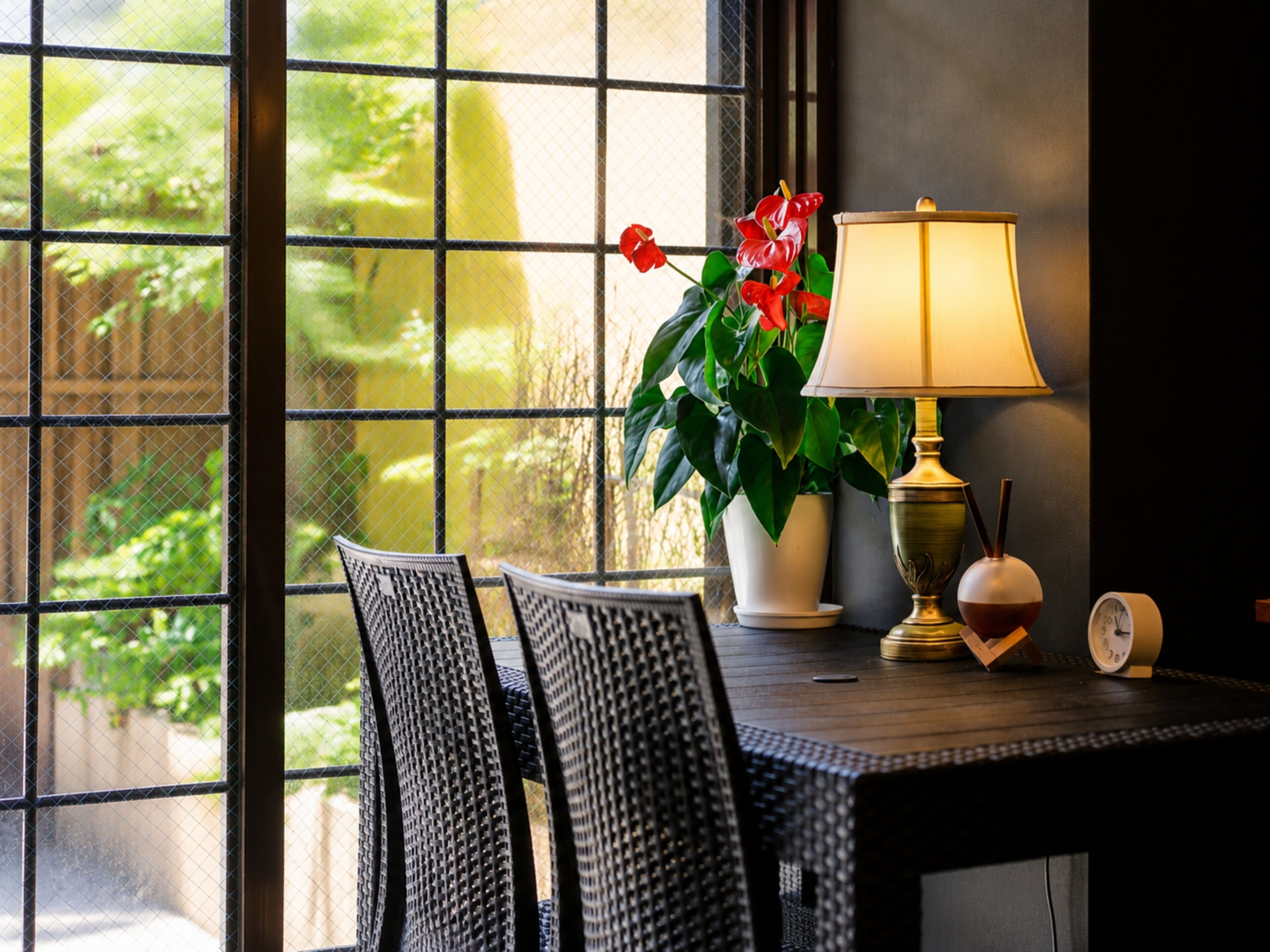 A room with a table, chairs, a lamp, a clock, and a window with a view of greenery.