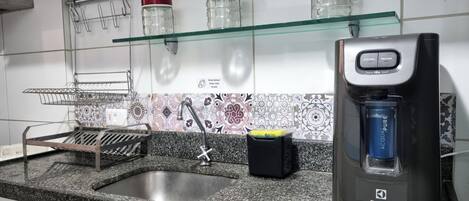A kitchen with a granite countertop, a stainless steel sink, a glass shelf with jars, a coffee maker, and a backsplash with patterned tiles.