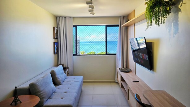 A modern living room with a grey sofa, a small round table, a TV mounted on the wall, and a window with a view of the sea. - Vista Mar na Pajuçara: Amplo 2Q p/ 6 Pessoas (Maceió)