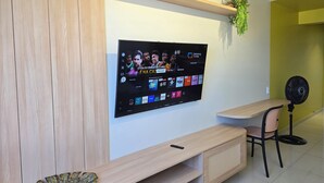 A modern living room with a flat-screen TV mounted on a wall, a wooden entertainment unit below, a small table with a chair, and a fan on the floor. - Vista Mar na Pajuçara: Amplo 2Q p/ 6 Pessoas (Maceió)
