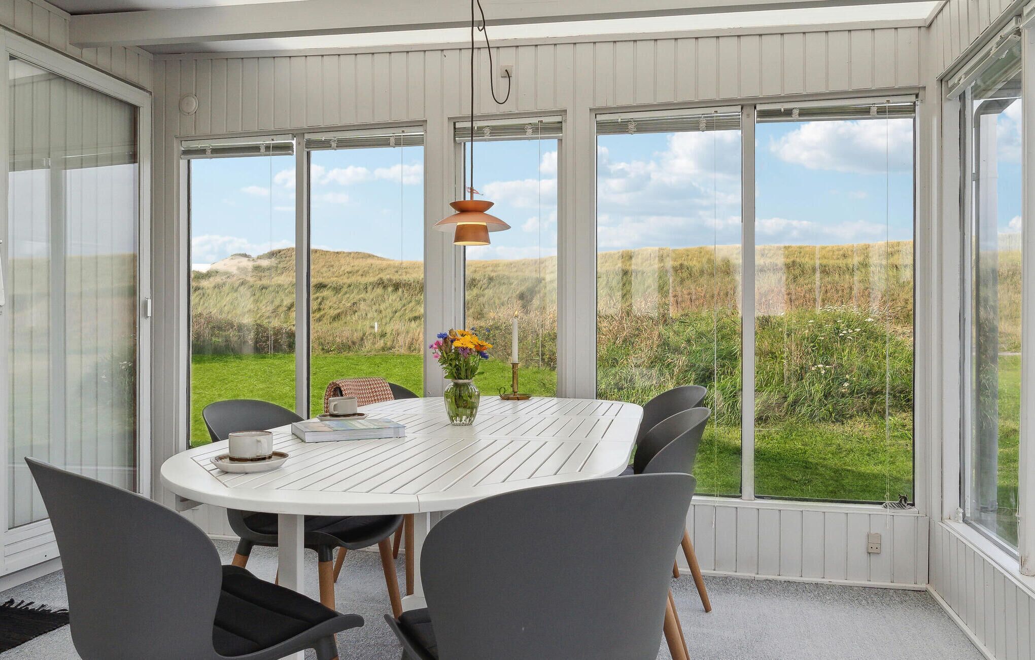 A dining area with a white table and grey chairs, large windows offering a view of a grassy landscape.
