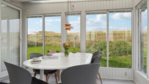Un espace repas avec une table blanche et des chaises grises, de grandes fenêtres donnant sur un paysage verdoyant.