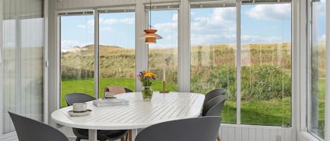 A dining area with a white table and grey chairs, large windows offering a view of a grassy landscape.