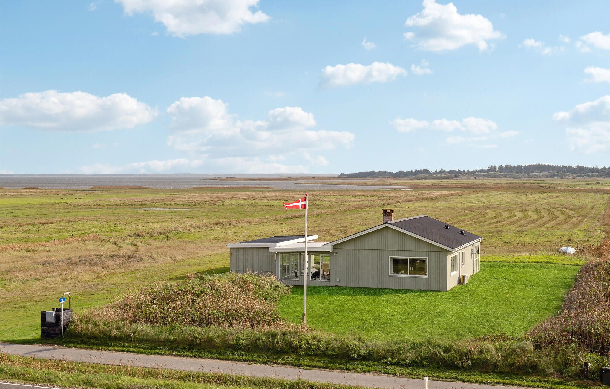 A small, single-story building with a flagpole, situated in a grassy field with a view of a distant shoreline.