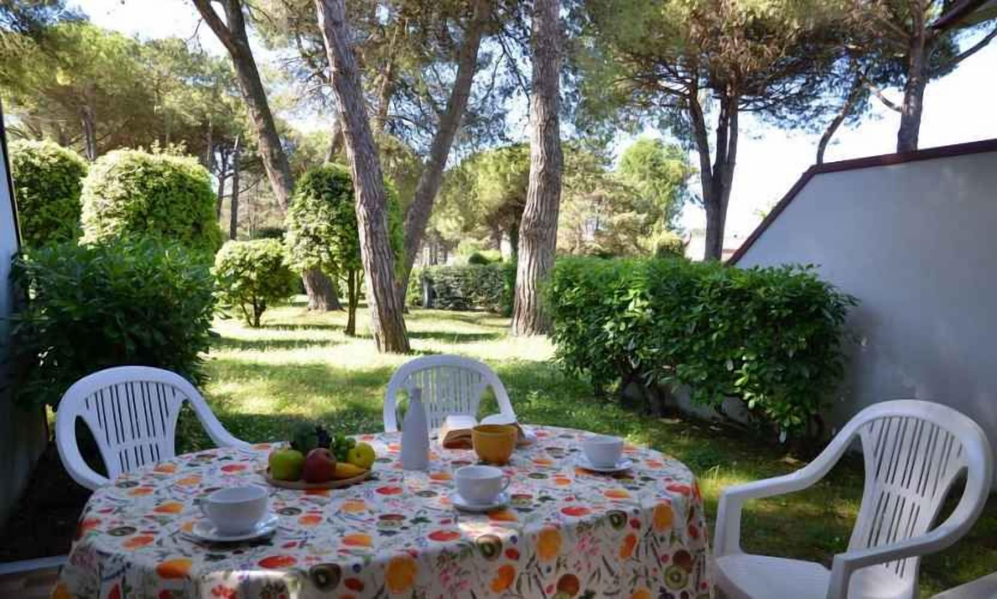 A round table with chairs and a fruit bowl, surrounded by greenery and trees.