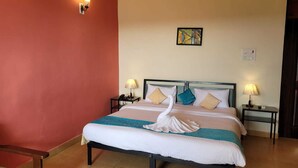 A neatly made bed with a white duvet, teal and beige pillows, and a white towel folded at the foot. There are two bedside tables with lamps, a wall-mounted light, and a small painting above the bed. - Palmarinha De Goa unit of GKP Real Estat (Calangute)
