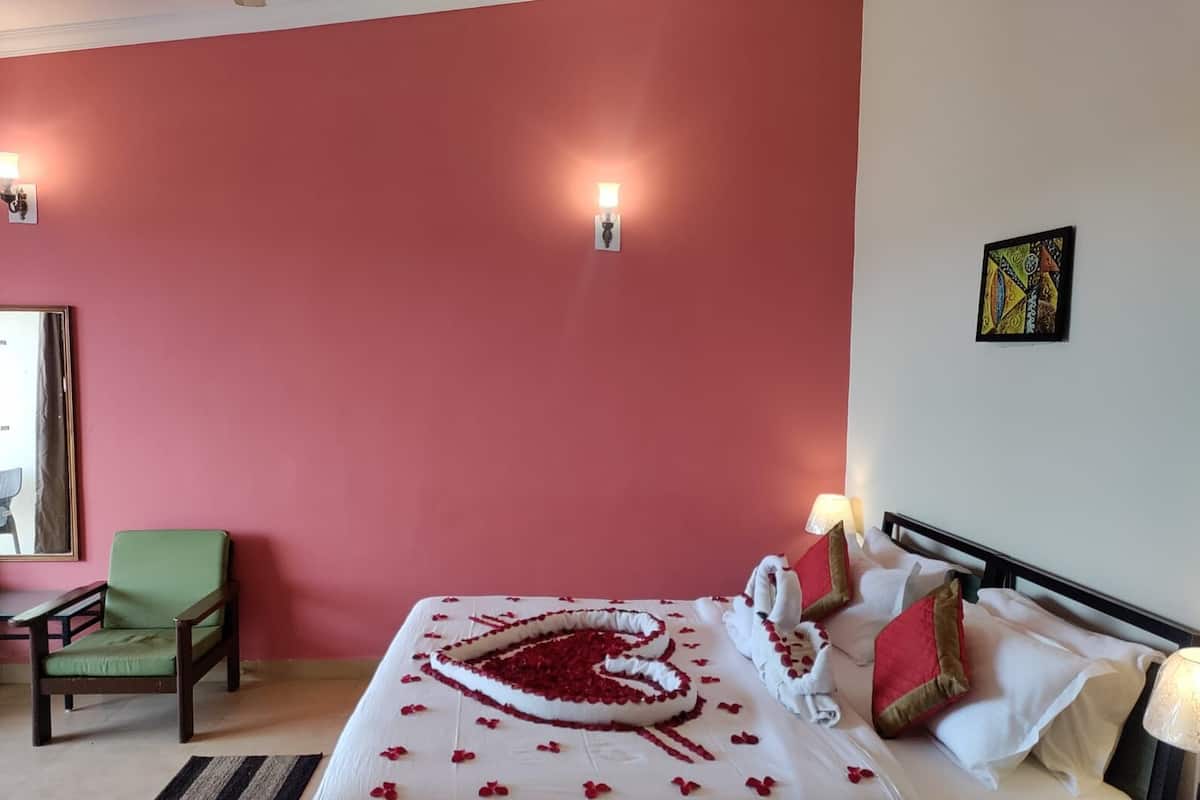 Ein Hotelzimmer mit roter Wand, einem Bett mit herzförmiger Dekoration, einem grünen Sessel und zwei Wandleuchten.