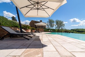 Ein Poolbereich mit Liegestühlen und Sonnenschirmen, ein Backsteinhaus im Hintergrund und ein klarer, blauer Himmel.