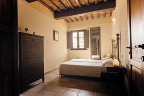 A bedroom with a wooden wardrobe, a bed with white bedding, a nightstand with a candle, and a window with wooden shutters.
