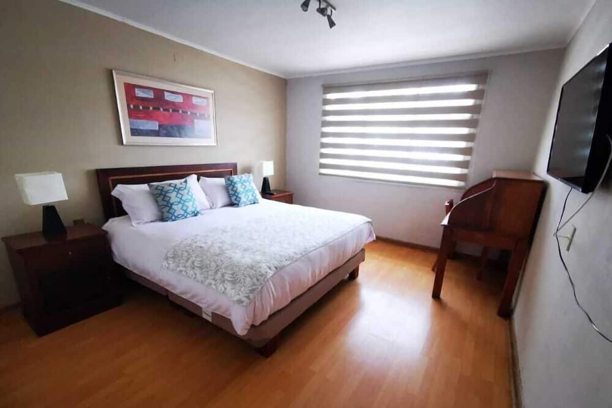 A bedroom with a bed, a nightstand, a lamp, a TV, and a window with blinds.