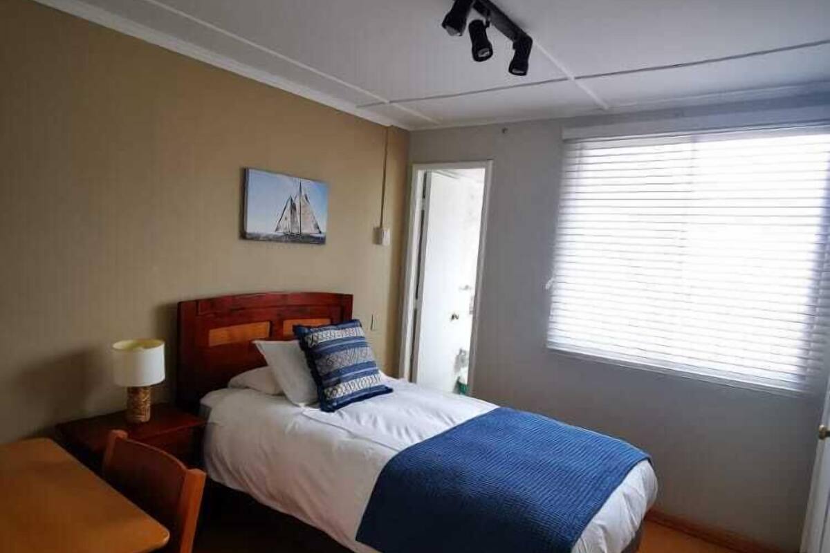 A bedroom with a bed, a nightstand, a chair, a table, a window with blinds, and a painting on the wall.