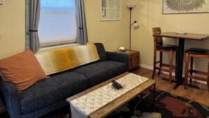 A living room with a dark blue sofa, a wooden coffee table, a small wooden side table, a lamp, and a dining area with two chairs. - Historic Townsend Park bungalow minutes from Black Canyon National Park (Montrose)