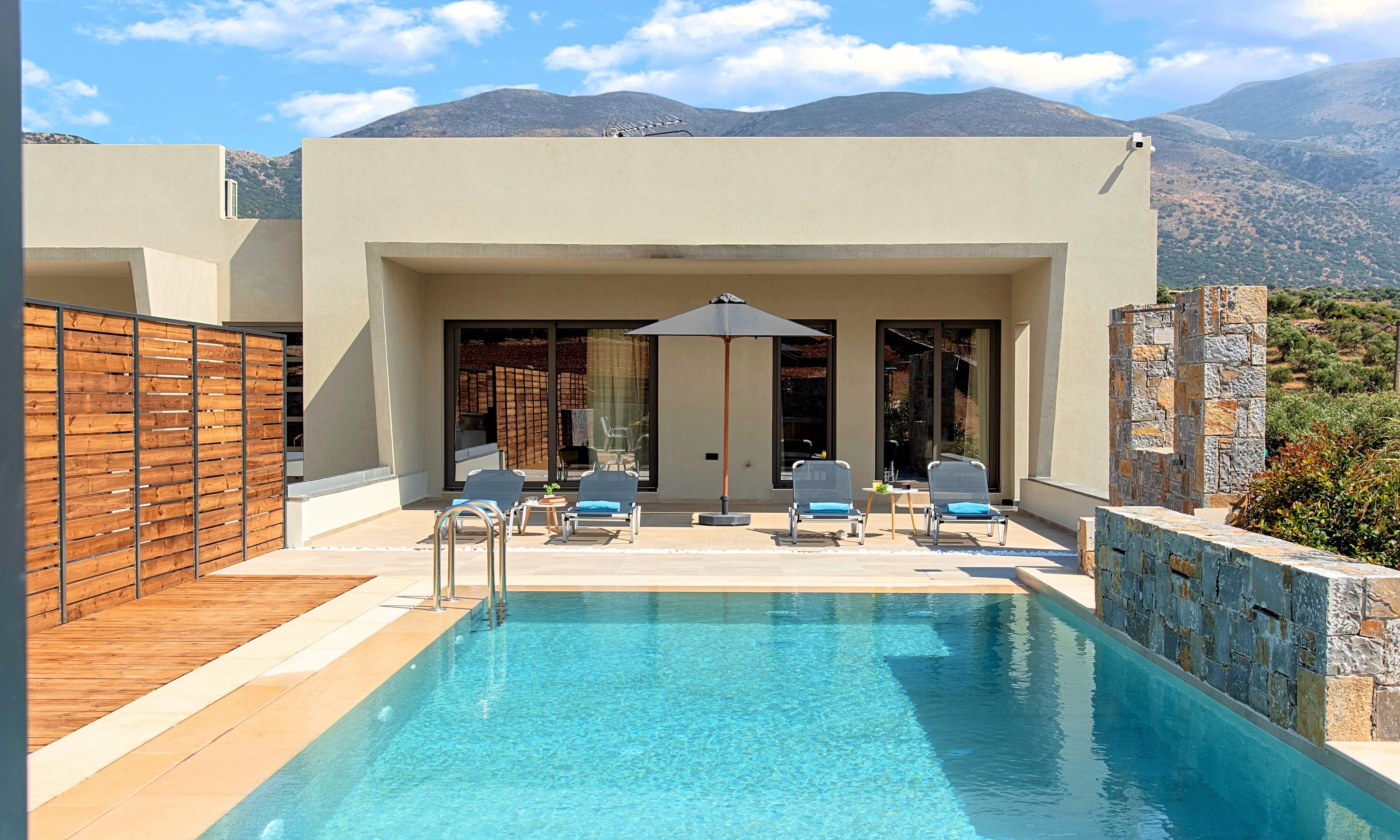 A pool area with lounge chairs and a table, surrounded by a wooden fence and stone walls, with mountains in the background.