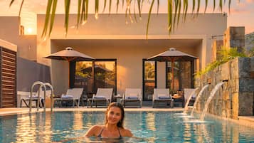 A swimming pool with lounge chairs and umbrellas, a waterfall feature, and a modern building in the background.