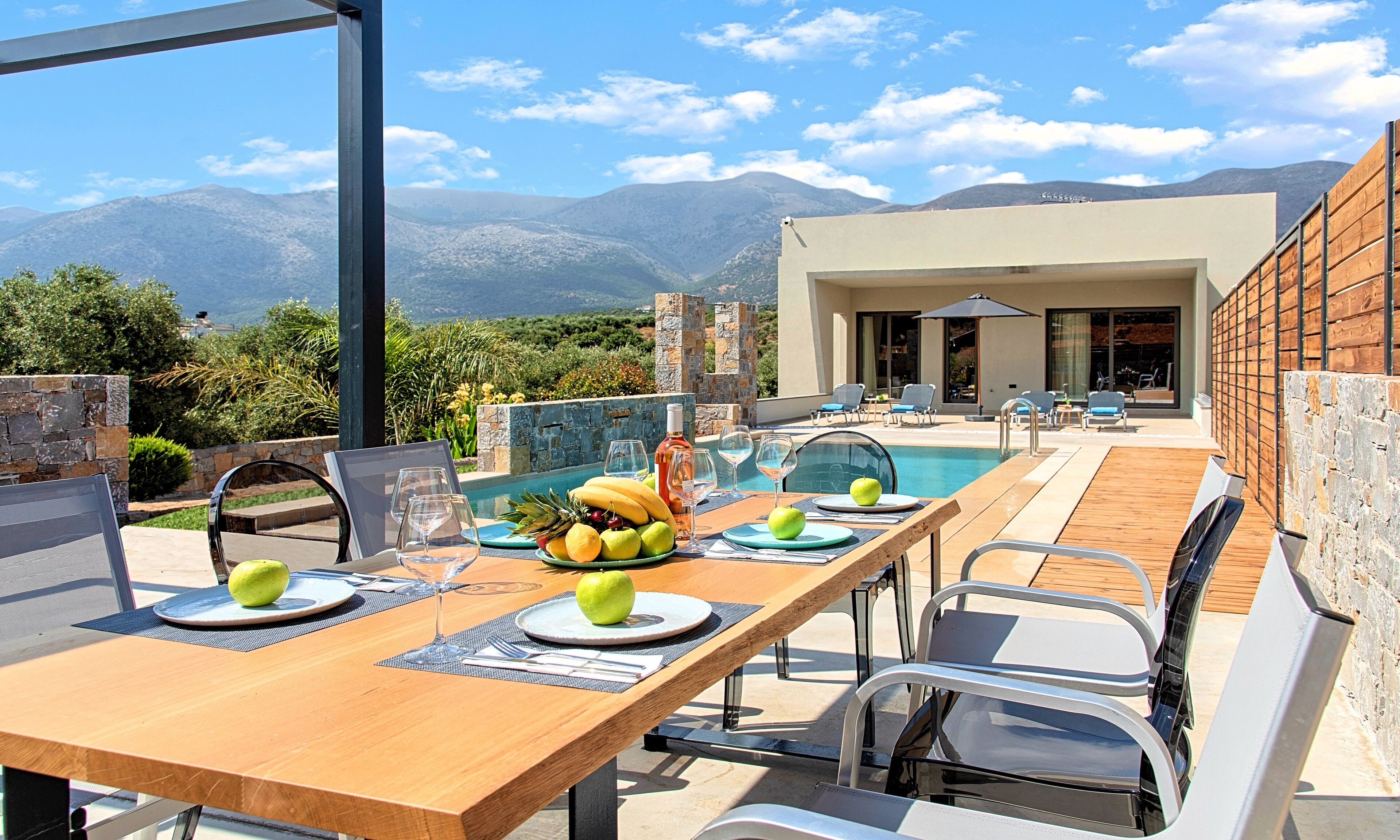 Un área de comedor al aire libre con piscina, vista a la montaña y un edificio moderno al fondo.