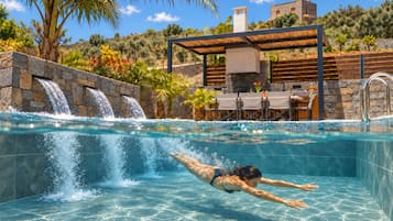 A swimming pool with a waterfall feature, a covered seating area, and a stone wall.