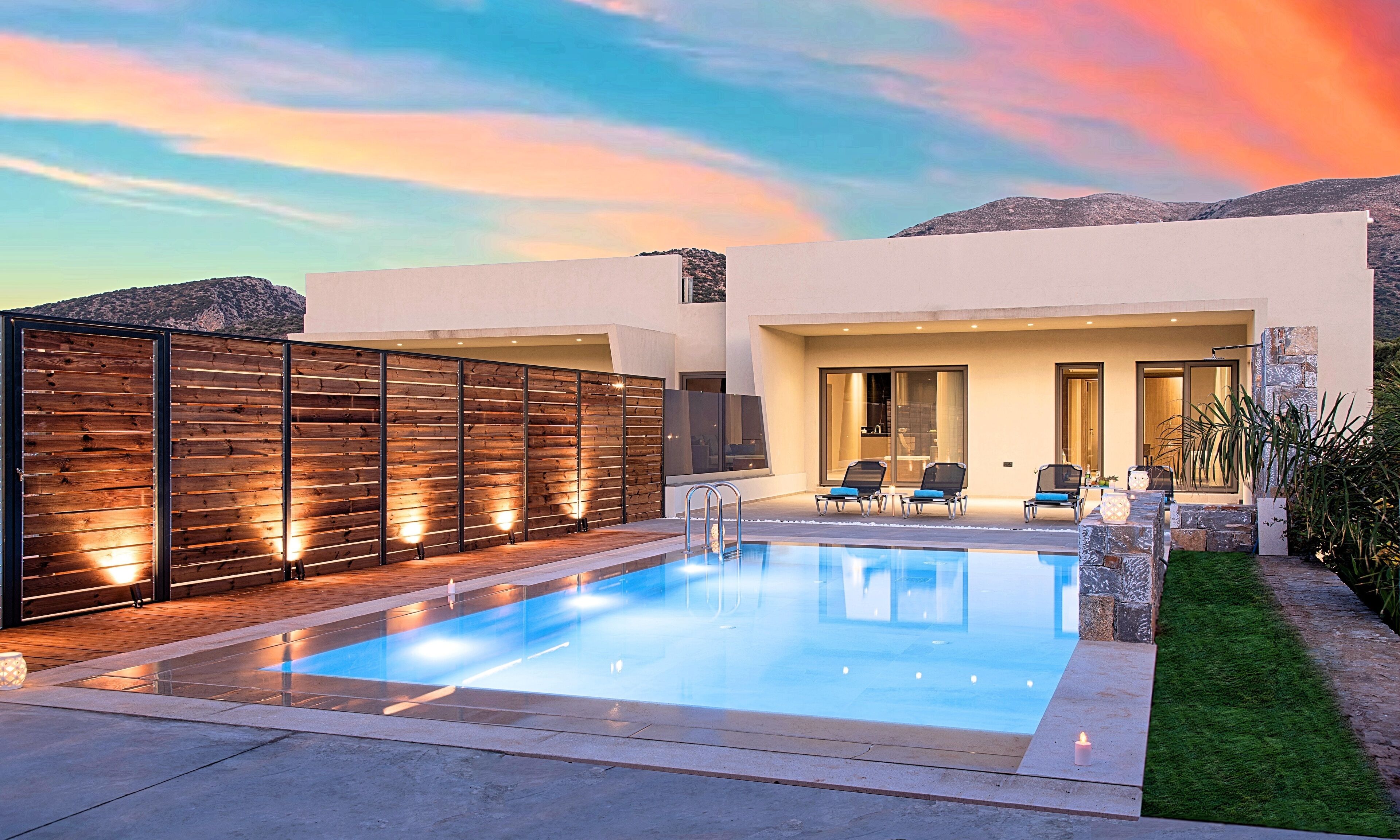 A modern outdoor pool area with a wooden fence, a seating area with chairs, and a view of the mountains in the background.