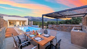 Un espace repas aménagé au bord de la piscine, avec une table dressée pour quatre personnes, entouré de murs en pierre et offrant une vue sur les montagnes.