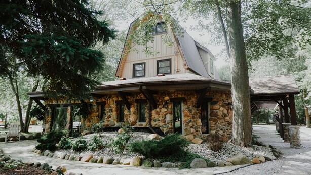 A stone house with a sloped roof, surrounded by a garden with various plants and a stone pathway.