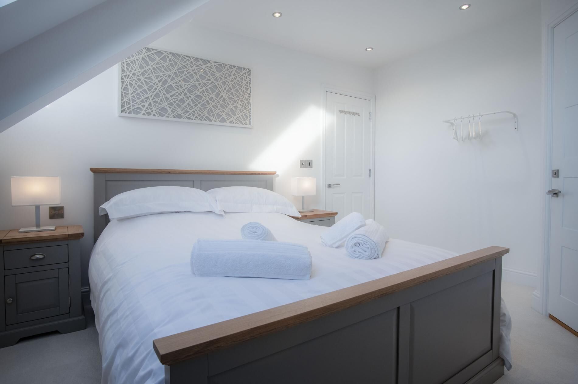 A bedroom with a bed, two bedside tables with lamps, a wooden headboard, a white door, and a wall-mounted towel rack.