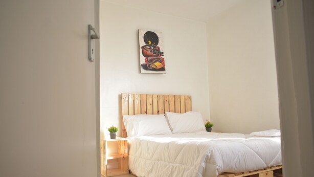 A bedroom with a wooden bed frame, white bedding, and a small bedside table with a plant. - Cozy stay in Langata (Nairobi)