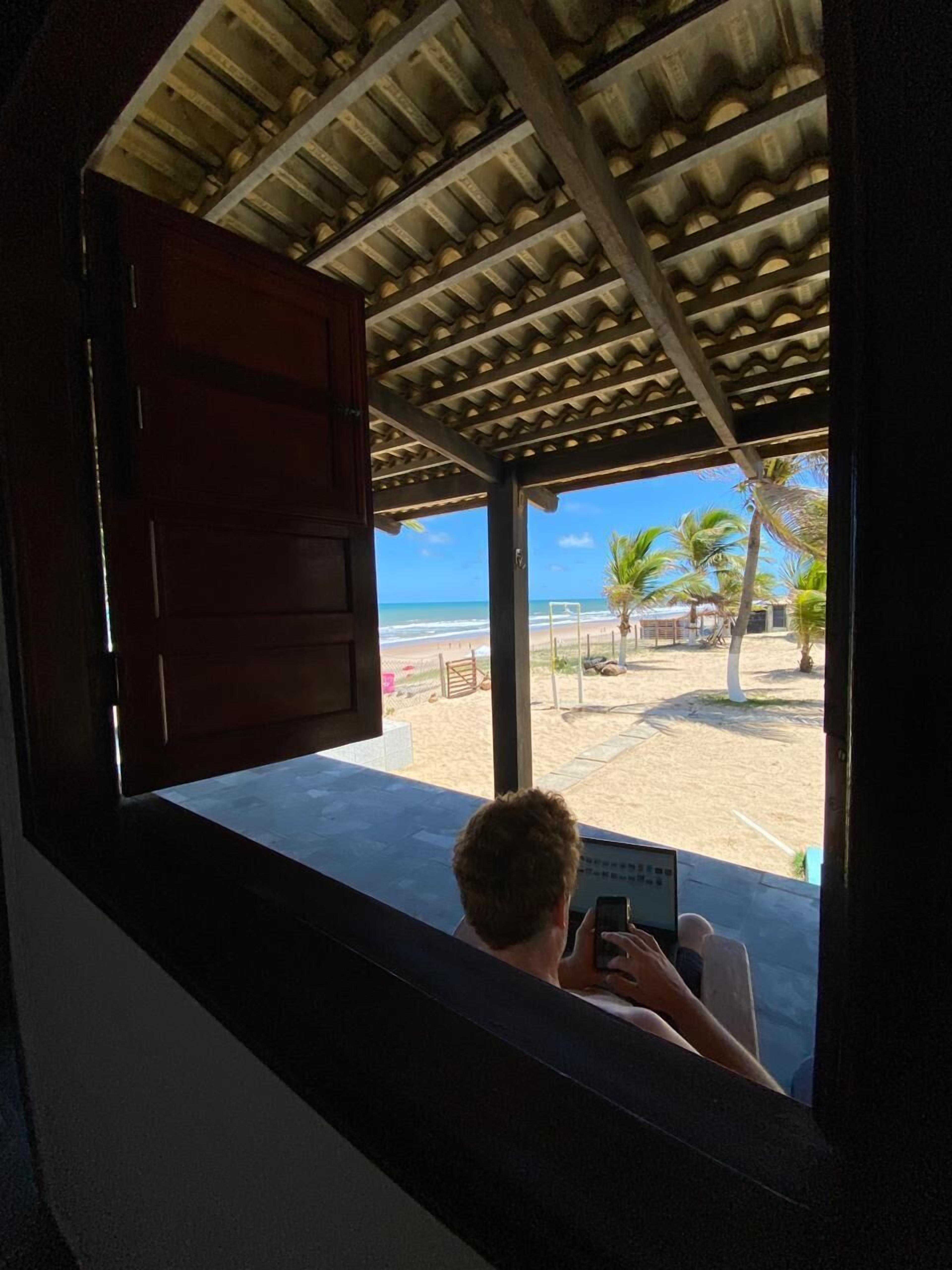 Una persona sentada junto a una ventana con vista a una playa y palmeras.