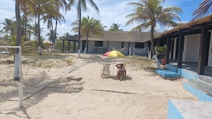 A sandy beach area with palm trees, a covered seating area, and a building with a tiled roof. - Pousada Marinheiro (Conde)