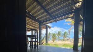 An open-air covered patio with a tiled floor and a floral rug, leading to a view of palm trees and a beach. - Pousada Marinheiro (Conde)