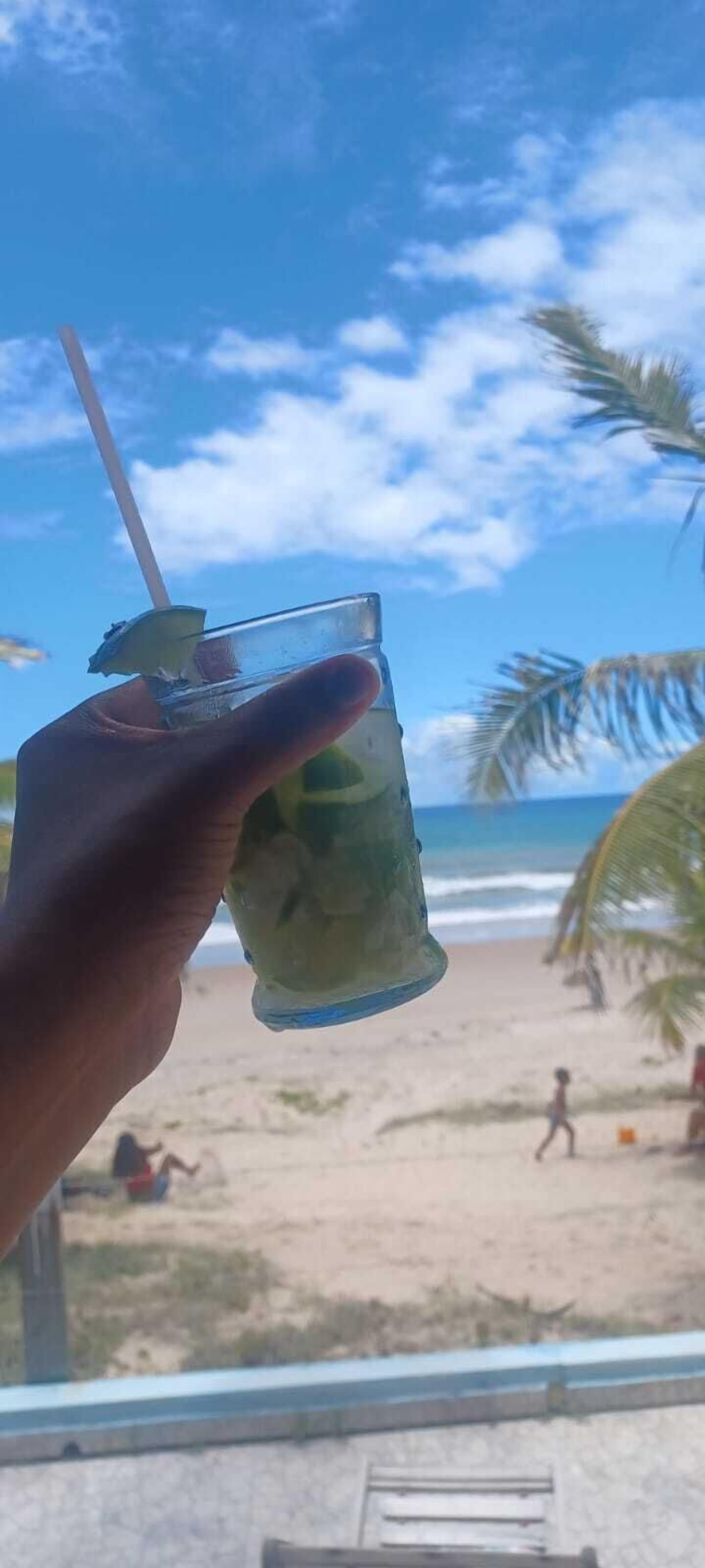 Una mano sosteniendo un vaso con pajita, con una playa y palmeras al fondo.