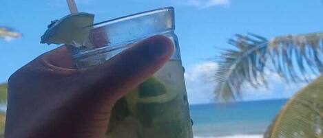 Una mano sosteniendo un vaso con pajita, con una playa y palmeras al fondo.