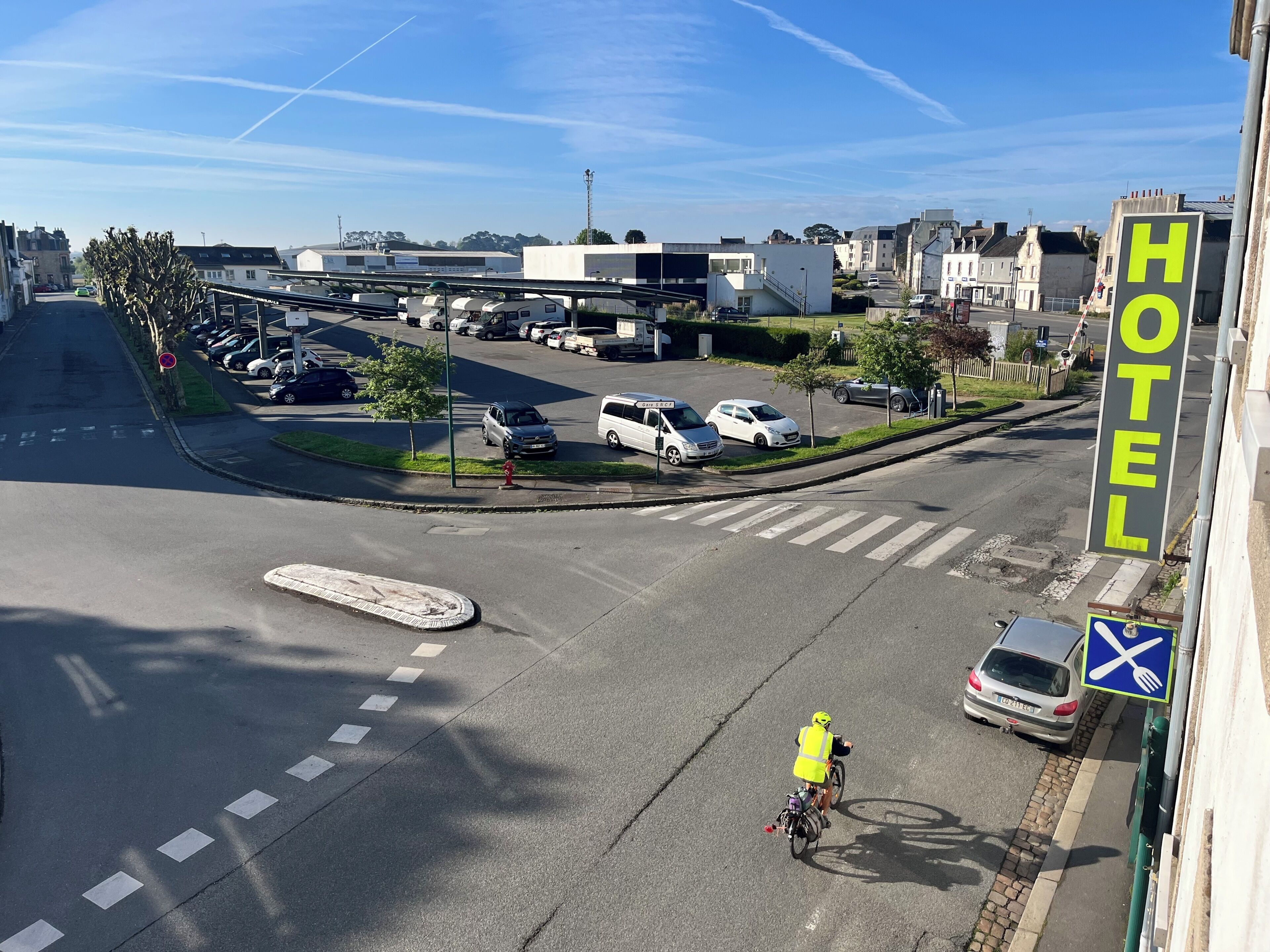 Une enseigne d’hôtel, un cycliste et un parking avec des voitures et un passage piéton.
