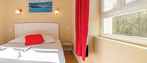 A hotel room with a bed, a nightstand, a small table with a plant, a painting on the wall, and a window with red curtains.