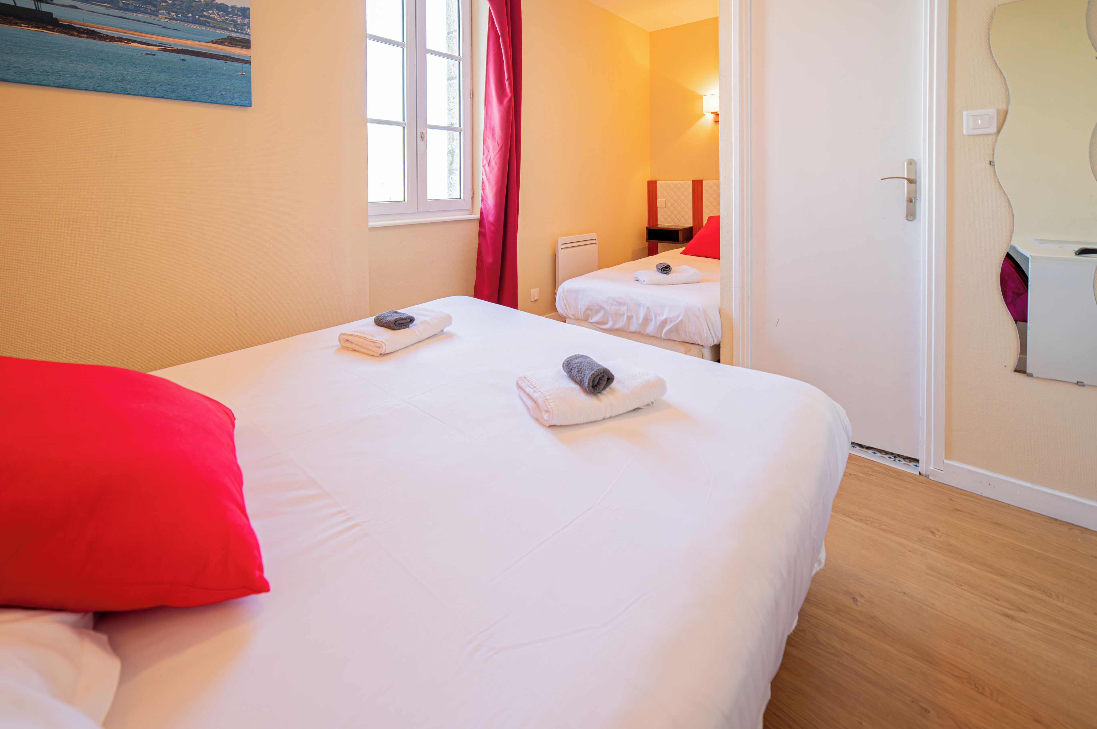 Une chambre d’hôtel avec deux lits, chacun agrémenté d’une serviette roulée, d’un oreiller rouge et d’un tableau représentant une plage, accroché au mur.