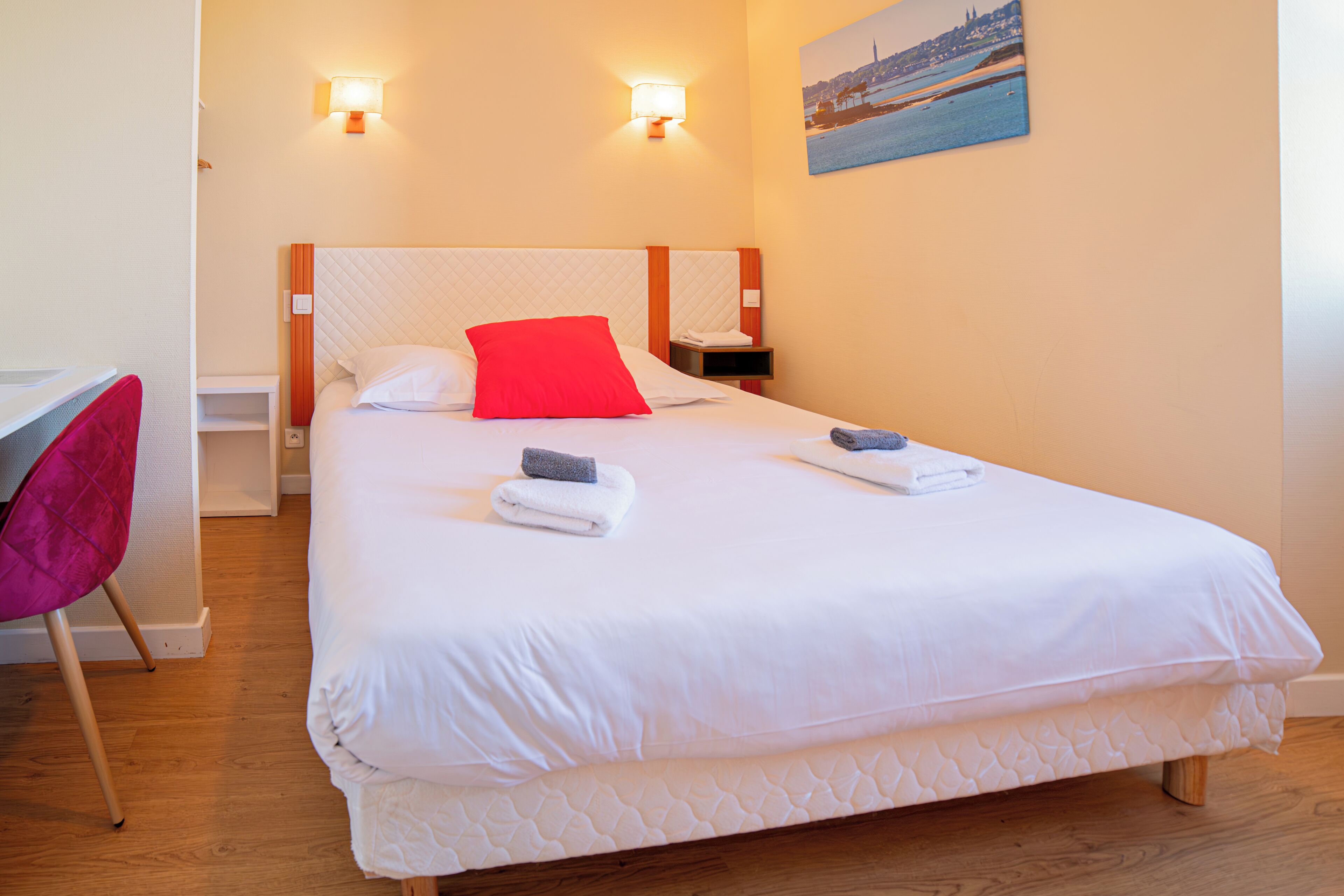 Una cama bien tendida con ropa blanca y una almohada roja, una mesita de noche de madera con una lámpara, y una silla con un cojín rosa.