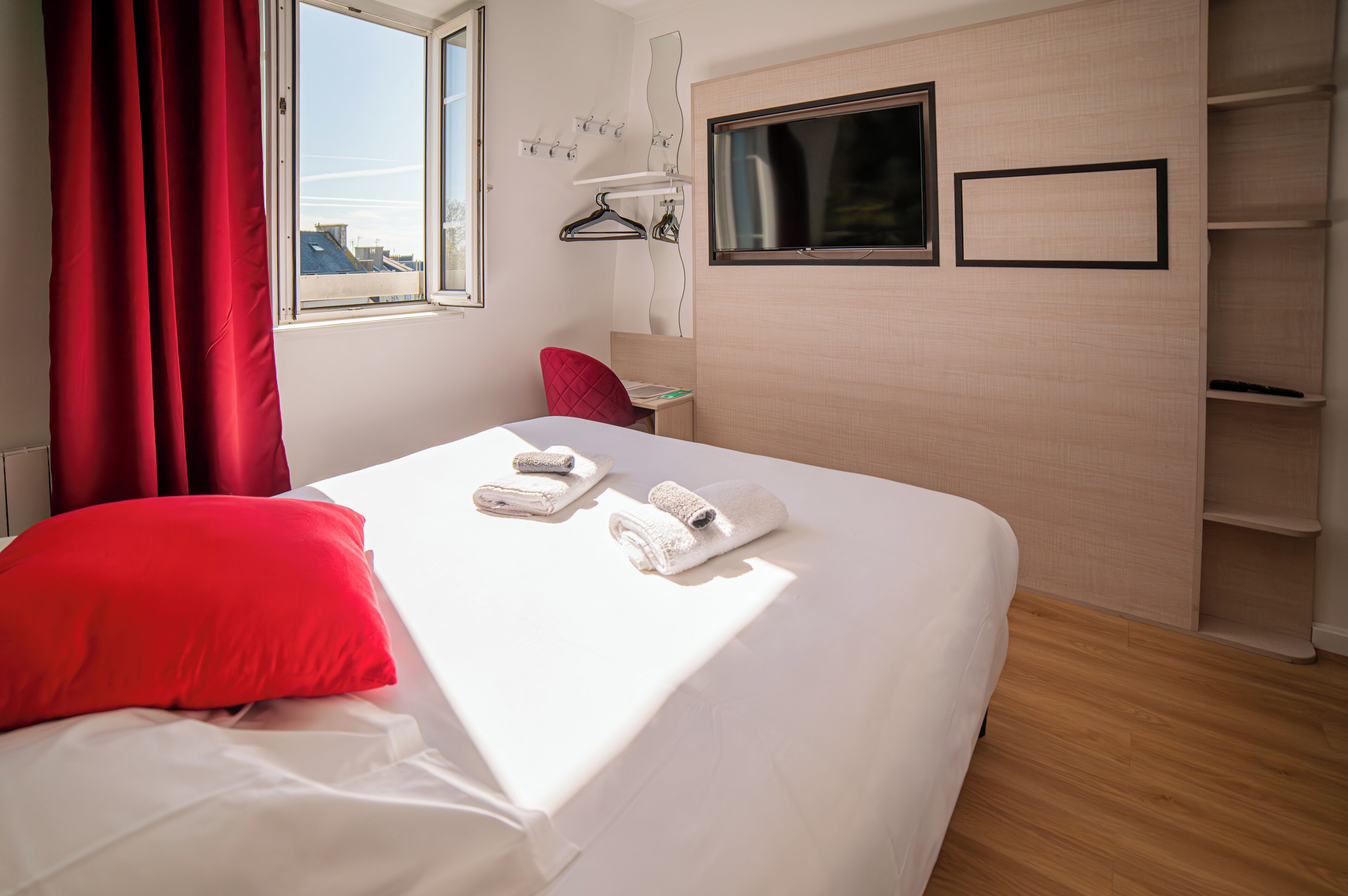 Una habitación de hotel con cama, cortinas rojas, una ventana, televisor y repisa mural.