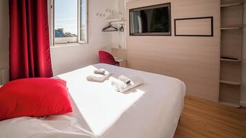 Una habitación de hotel con cama, cortinas rojas, una ventana, televisor y repisa mural.