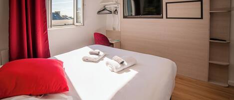 A hotel room with a bed, red curtains, a window, a TV, and a wall-mounted shelf.