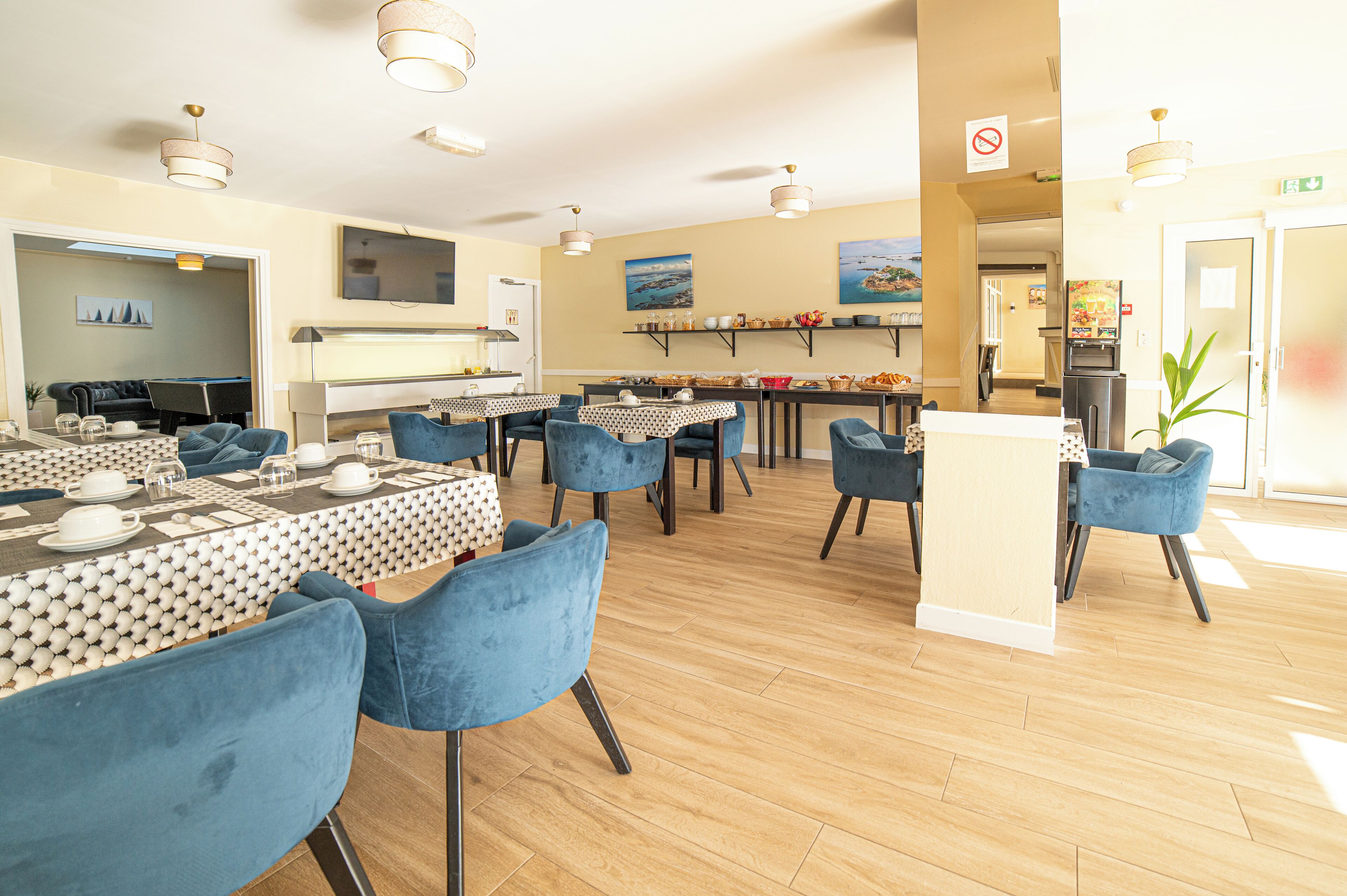 Un espace repas avec plusieurs tables et des chaises bleues, un piano et un téléviseur fixé au mur.