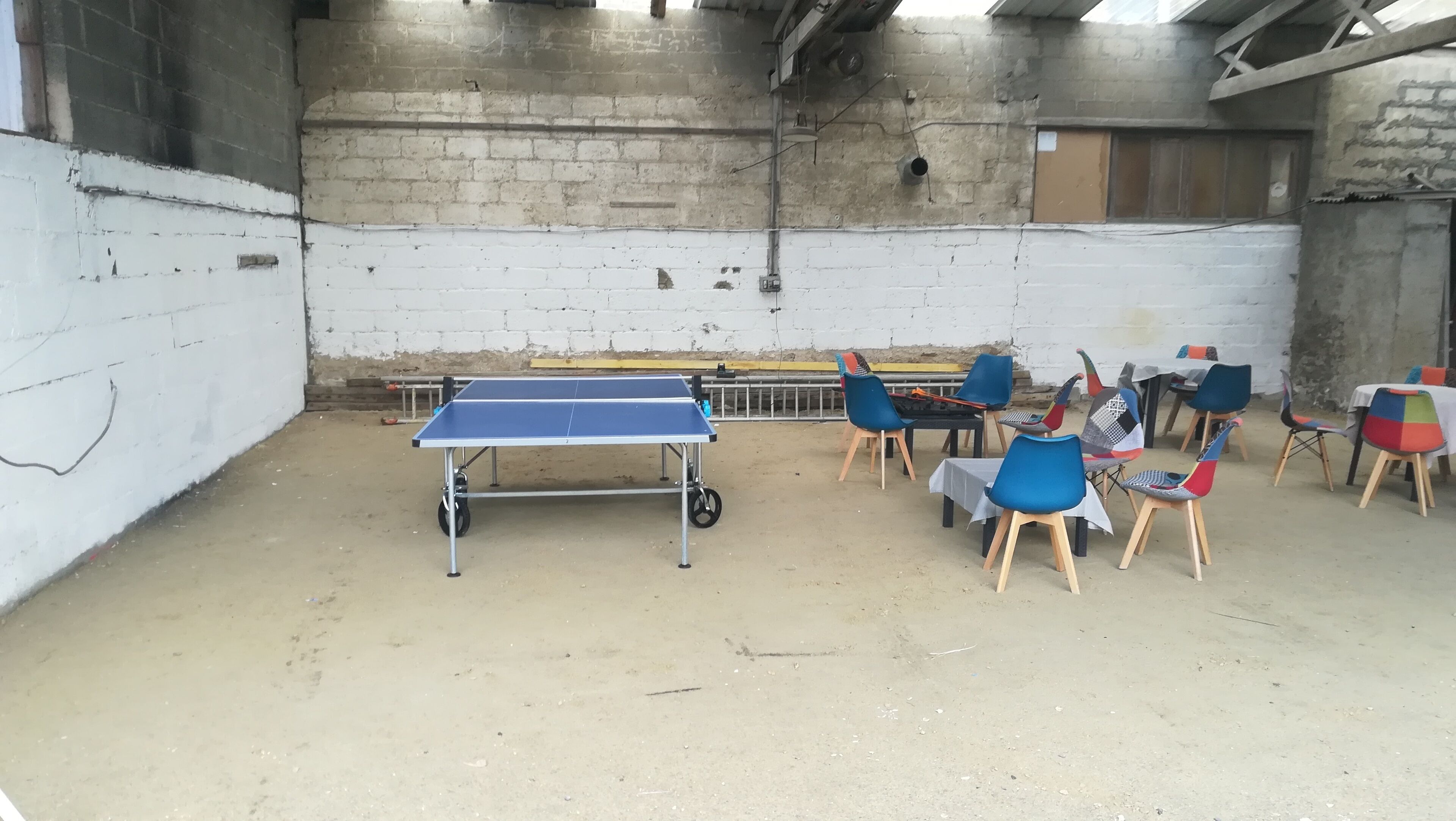A ping pong table in a room with white walls and a concrete floor. There are several chairs around the table.