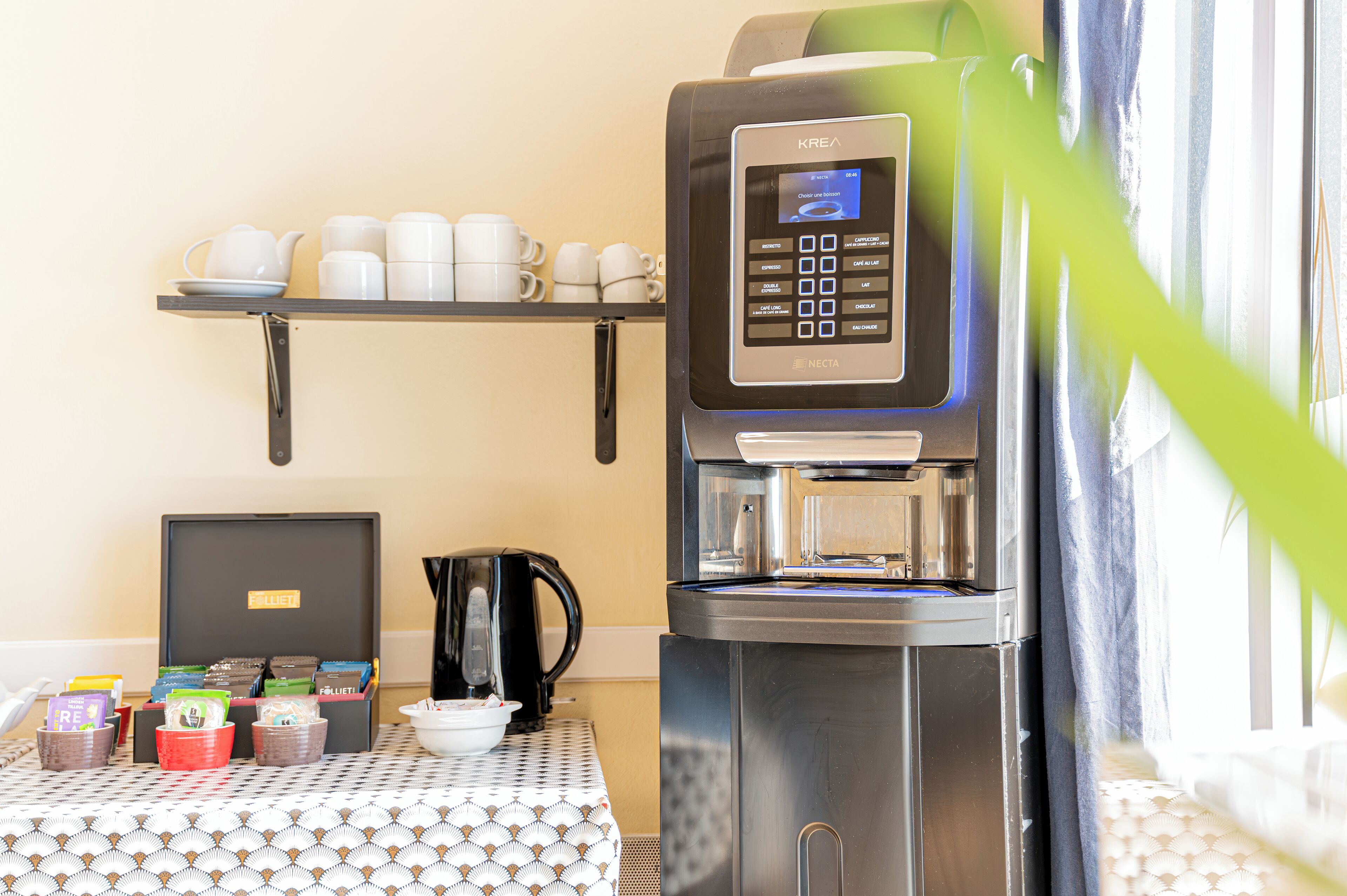 Een koffieapparaat met een digitaal display, een plank met bekers, een waterkoker en een tafel met voedselverpakkingen.